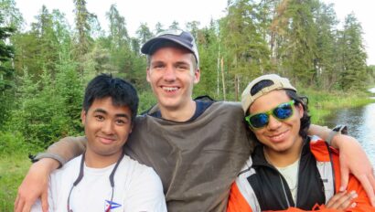 three friends smiling for the camera outdoors next to a lake.